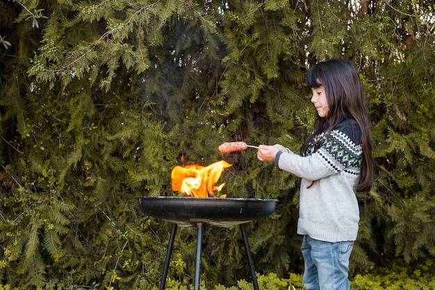 屋外のバーベキューの炎の上にソーセージを焼く少女