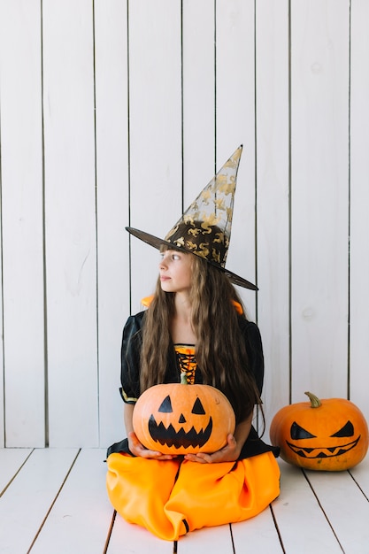 Free photo girl in halloween costume sitting and holding pumpkin in studio