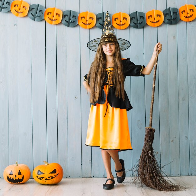 Girl in Halloween costume standing with broom and pumpkins