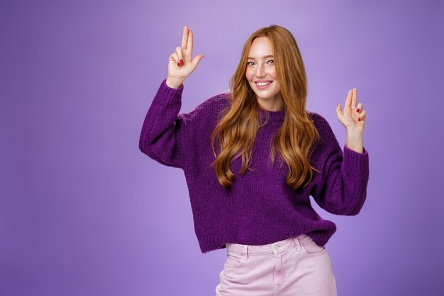 紫の壁の上の紫色のセーターでポーズをとる素晴らしいパーティーでスリルと喜びを感じてカメラで軽薄な笑顔のように指のピストルのジェスチャーを楽しんで楽しんで踊っている幸せで元気な気分の女の子。