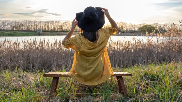 A girl in a hat and in casual style sits on a bench near the lake at sunset