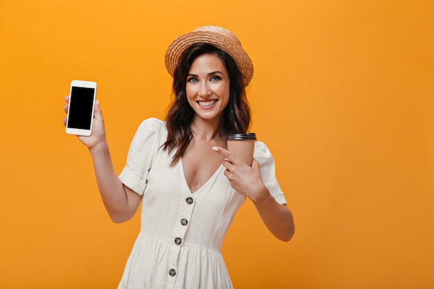 Ragazza in cappello e vestito mostra sullo smartphone e tiene un bicchiere di tè