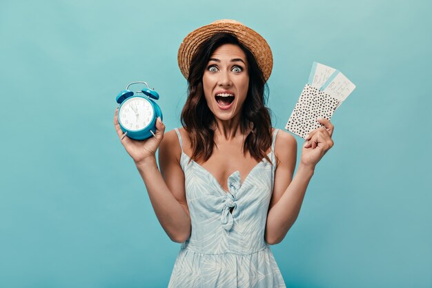 Girl in hat looks funny at camera and keeps alarm clock, tickets on blue background