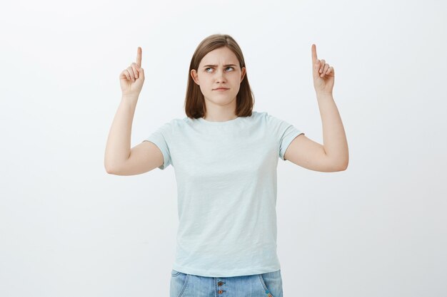 Girl have doubts think product is lame. Intense displeased cute girl with big ear frowning looking and pointing up with dislike and doubt hesitating if choice is right posing over white wall