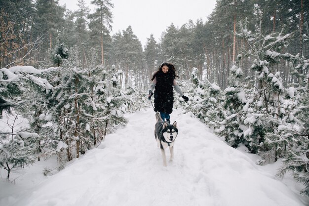 Девушка веселится с ее собакой Husky в снежном зимнем сосновом лесу