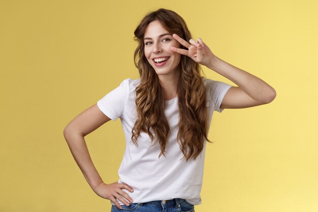 Girl having fun feel happy enthusiastic enjoy perfect summer day share positivity upbeat mood show peace victory sign near cheek tilt head smiling silly carefree stand yellow background. Lifestyle.