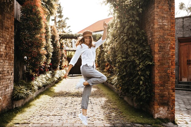 Girl in high spirits is jumping against space of old courtyard with ivy on fence. Snapshot of lady in white clothes.