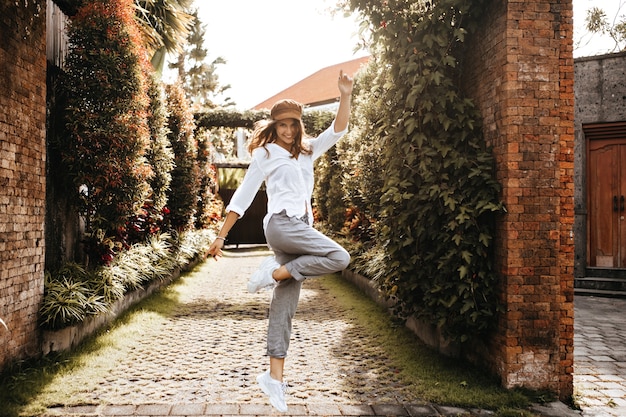 Girl in high spirits is jumping against space of old courtyard with ivy on fence. Snapshot of lady in white clothes.