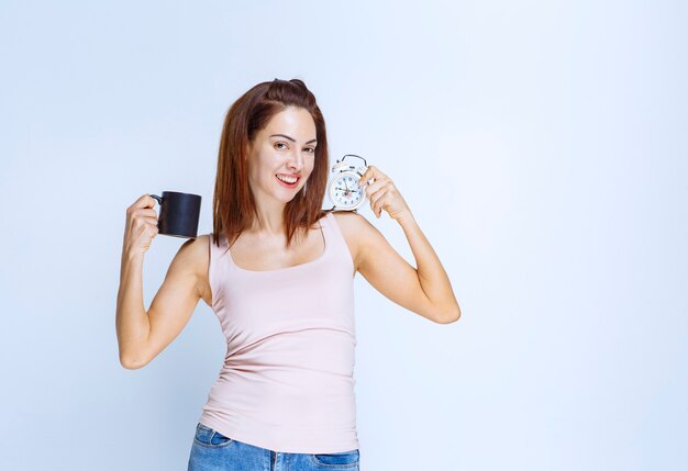 Girl holding an alarm clock in one hand and a black coffee mug in another. 