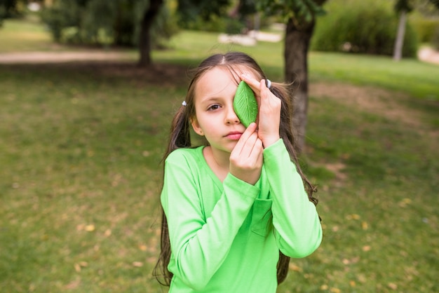 Foto gratuita ragazza che tiene foglia verde artificiale sul suo occhio sinistro in piedi sull'erba