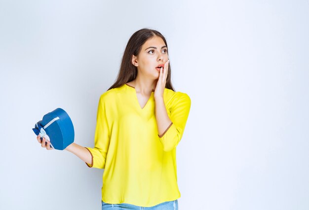 Girl holding a blue heart shape gift box and looking surprized and terrified.