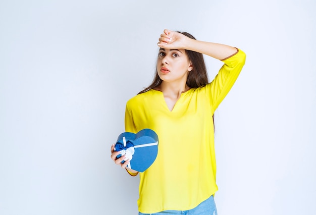 Girl holding a blue heart shape gift box and looks tired.