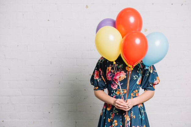 Girl holding colorful balloons in front of her face standing against wall