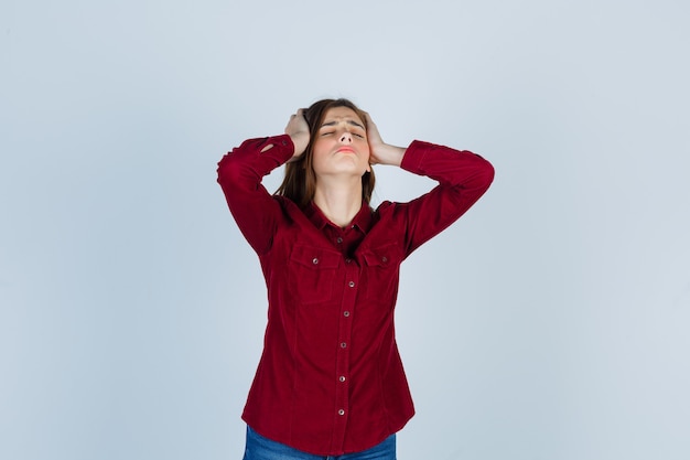 Ragazza che tiene le mani sulla testa in camicia bordeaux e sembra infastidita.