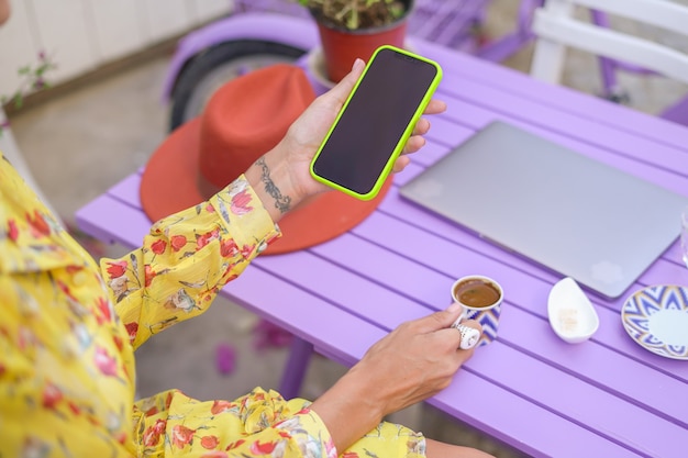 카페에서 빈 검은색 화면이 있는 휴대전화를 들고 있는 소녀, 노트북, 터키식 커피가 탁자 위에 있습니다