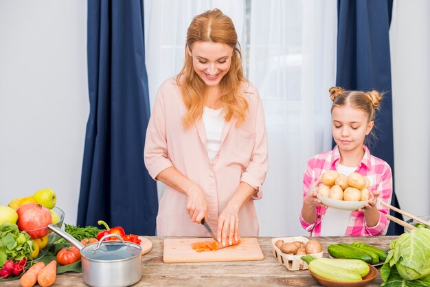Girl holding potatoes plate standing near the smiling young woman cutting the carrot with knife on table