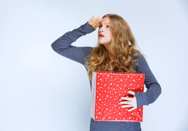 Девушка держит красную подарочную коробку и выглядит смущенной.