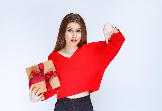 Girl holding a red ribbon wrapped cardboard gift box and inviting someone to receive it. 