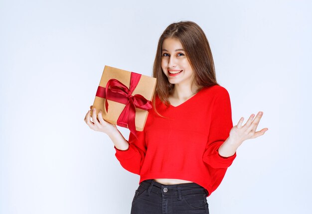 Девушка держит обернутую красной лентой картонную подарочную коробку и указывая на кого-то.
