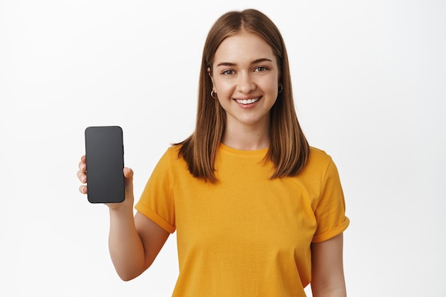 Girl holding smartphone and smiling, showing interface app, mobile phone empty screen, standing in yellow t-shirt over white wall.