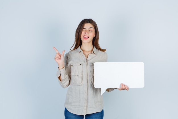 Free photo girl holding speech bubble, pointing at upper left corner while blinking in shirt and looking happy