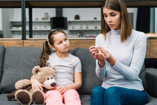 Girl holding teddy bear sitting near the young woman looking at temperature