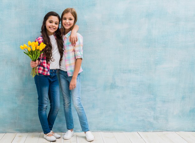 Девушка держа тюльпаны в руке стоя с ее подругой против голубой стены