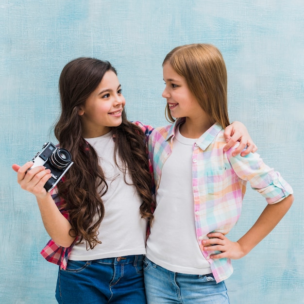 Girl holding vintage camera in hand looking at her female friend against blue wall