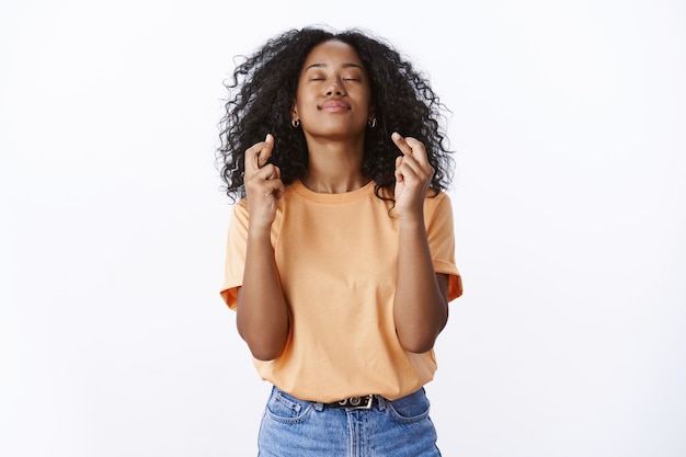 Girl hopes god hears prayers making wish desire come true. Hopeful attractive young female student wishes pass university exams close eyes raise head sky, cross fingers good luck white wall