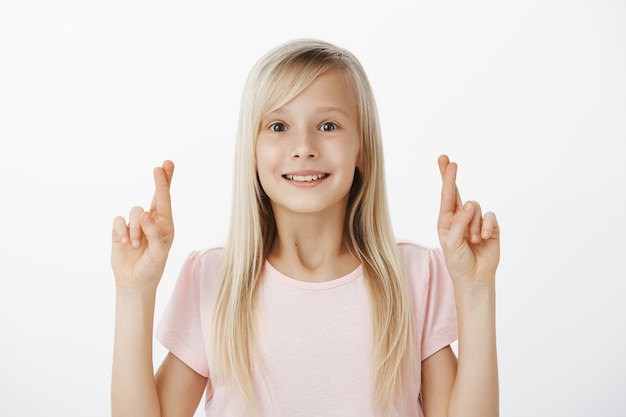 Girl hopes santa will visit her house with huge present. Grinning dreamy young daughter with blond hair, raising hands with crossed fingers and smiling with wonder and desire, hoping or making wish