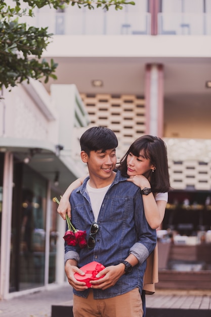 Girl hugging a man from behind and this holds roses and a gift