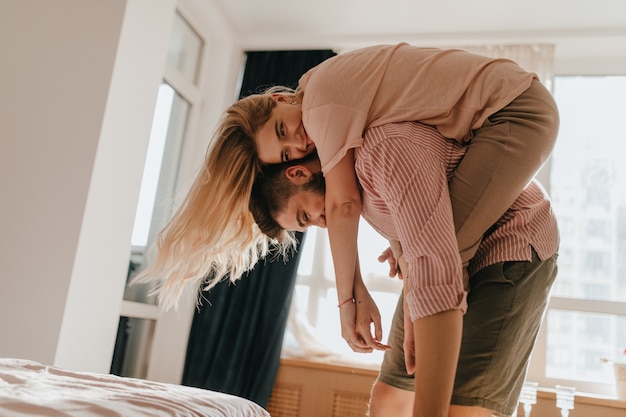 Free photo girl hung on back of her beloved boyfriend and smiles. young married couple are naughty and have good time in their bright, cozy bedroom.