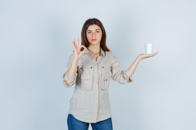 Девушка в рубашке держит пластиковую чашку кофе, показывает знак ок и выглядит довольной.