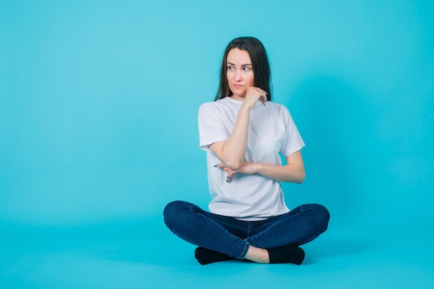 Girl is looking away by holding hand on chin and sitting on floor on blue background