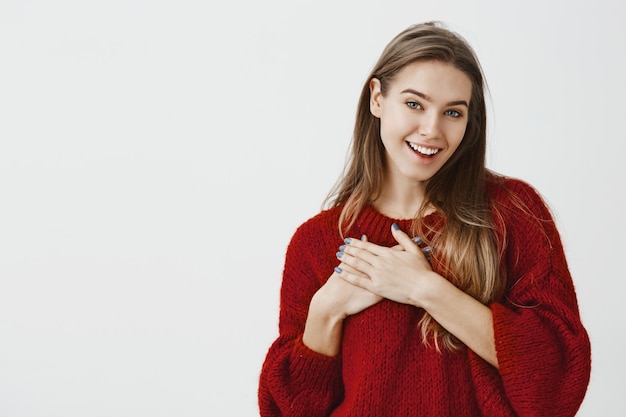 Бесплатное фото Девушка довольна комплиментами от corowker в офисе. тронутая очаровательная европейская женская модель в стильном красном свободном свитере, держащая ладонь на груди и улыбающаяся от удовлетворения над серой стеной
