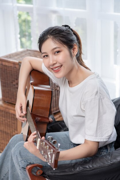 The girl is sitting and playing the guitar on the chair.