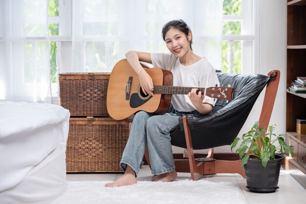 The girl is sitting and playing the guitar on the chair.