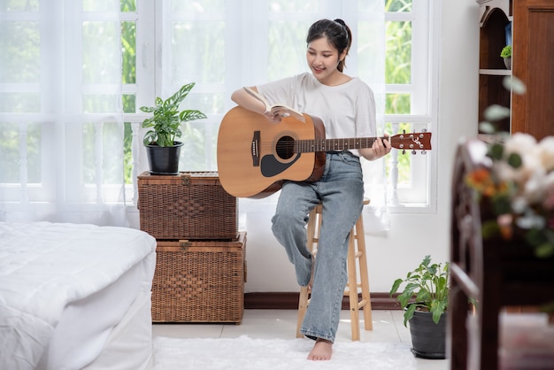 The girl is sitting and playing the guitar on the chair.