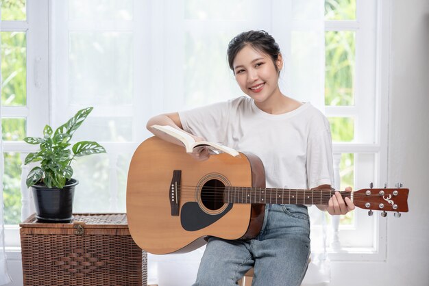 The girl is sitting and playing the guitar on the chair.