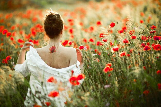 Girl is taking off the shirt from her back with a tattoo flower poppy on it, among the poppies field