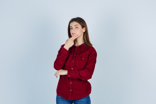 Девушка в бордовой блузке держит пальцы на подбородке и выглядит уверенно