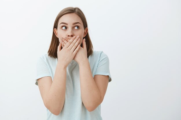 Girl learn shocking secret trying keep it. Portrait of shocked and astonished concerned speechless woman covering mouth with both palms gazing worried at upper right corner over grey wall