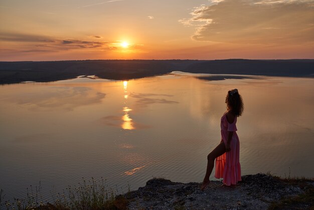 湖の近くの夕日を背景に明るいドレスの女の子