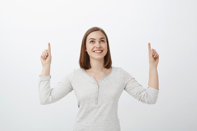 Girl likes counting start. Portrait of charming dreamy young woman with short brown haircut smiling joyfully looking and pointing up while enjoying great summer night posing against grey wall