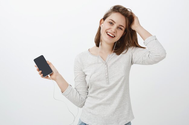 Girl likes working with earphones in ears. Joyful cute carefree european female touching hair gently tilting head, smiling joyfully listening music in earbuds holding smartphone showing gadget screen