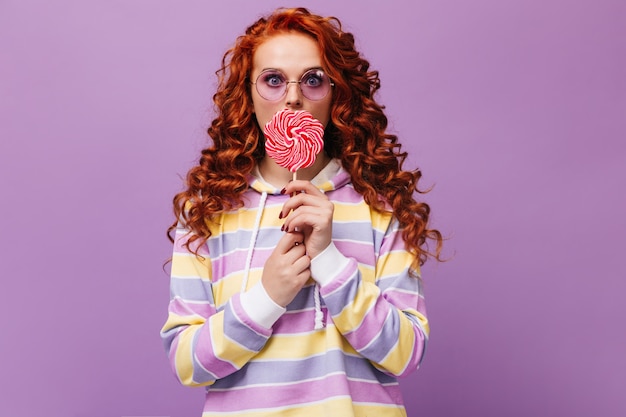 Free photo girl in lilac glasses and cute sweatshirt licks huge caramel and looks at front
