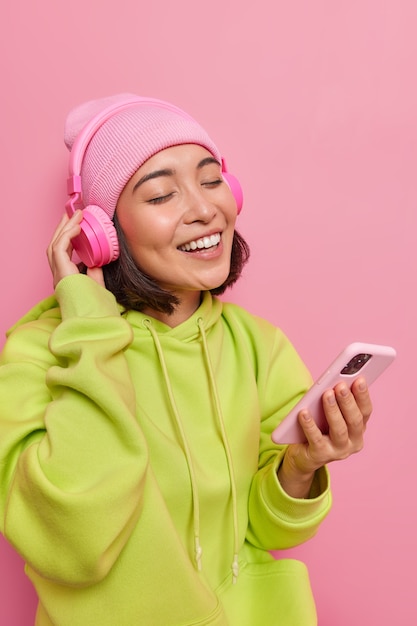 girl listens favorite music keeps eyes closed from pleasure holds mobile phone wireless headphones on ears dressed in green sweatshirt and hat poses on pink wall