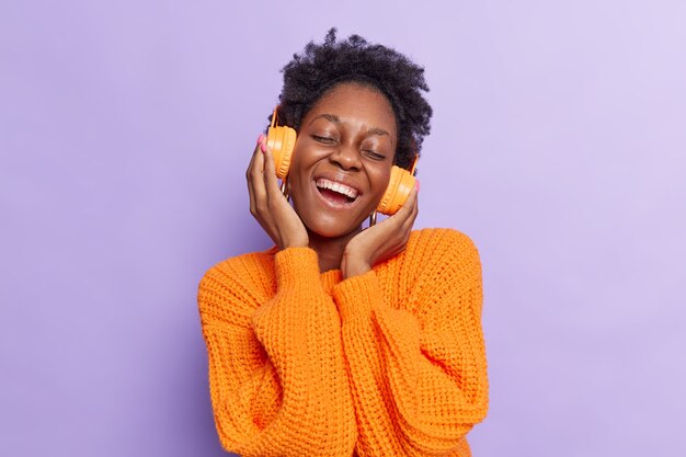 girl listens music via wireless headphones has fun dressed in casual knitted jumper isolated on purple 