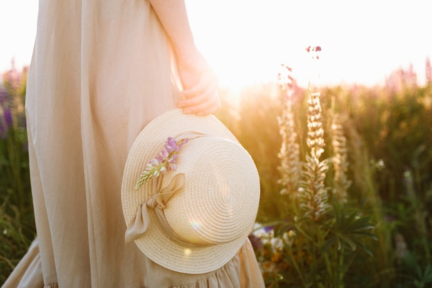 girl in long dress holding straw hat standing in spring flower field at sunset