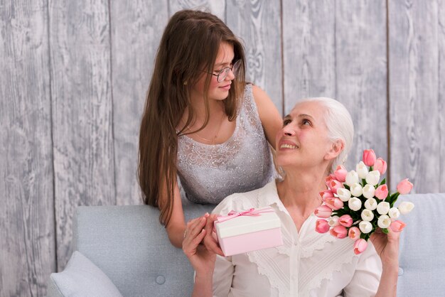 Girl looking at her grandmother holding gift box and tulip flower bouquet in hands